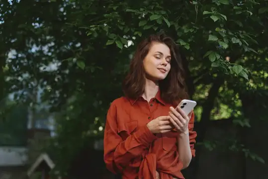 Молодая женщина в красной рубашке со смартфоном в руках среди зелени