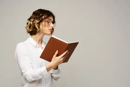 Молодая женщина в очках читает книгу в коричневом переплете