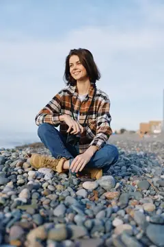 Улыбающаяся девушка с фотокамерой сидит на каменистом пляже
