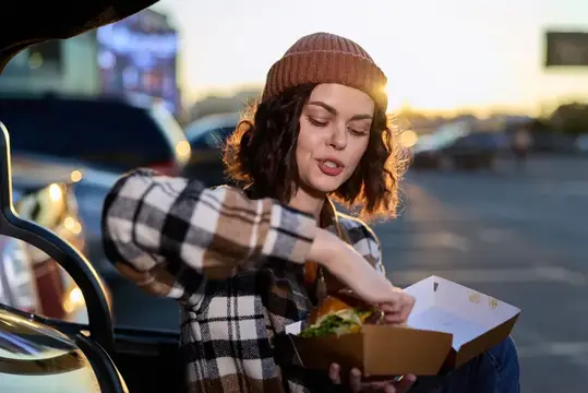 Девушка в шапке ест буррито у машины на закате