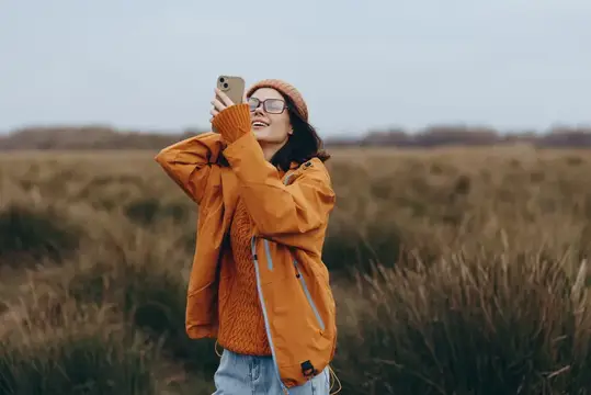 Улыбающаяся девушка в оранжевой куртке фотографирует на телефон в поле