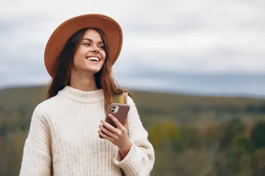 Улыбающаяся девушка в шляпе со смартфоном на фоне холмов