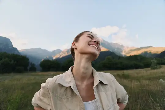 Молодая женщина смеётся с закрытыми глазами на фоне гор