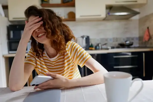 Кудрявая девушка смеётся, закрыв лицо рукой, с телефоном на кухне