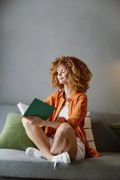 Рыжеволосая девушка с книгой сидит на диване с подушками