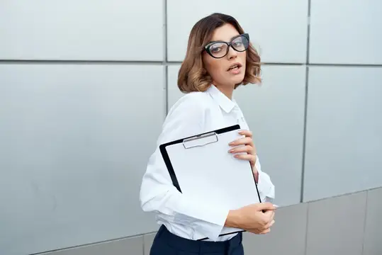 Удивлённая женщина в очках с планшетом у серой стены