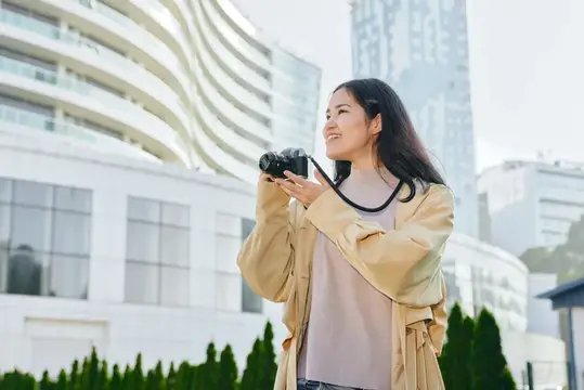 Улыбающаяся азиатская девушка с фотокамерой на фоне городских зданий
