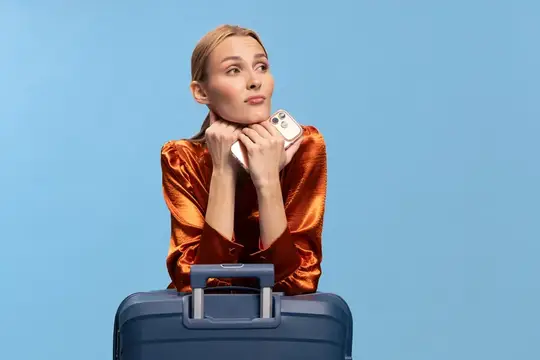 Задумчивая молодая путешественница в блестящем оранжевом топе держит телефон и отдыхает, опираясь на синий чемодан, изолированный на голубом фоне