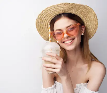 Улыбающаяся девушка в соломенной шляпе и очках пьёт напиток