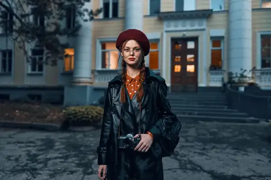 Рыжеволосая девушка в берете и кожаном плаще с фотокамерой на фоне старинного здания