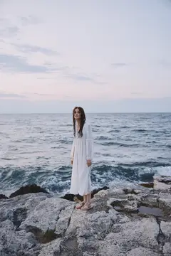 Девушка в белом платье стоит на скалах у моря