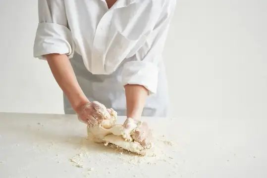 Женщина в поварской форме на кухне раскатывает тесто, занимаясь выпечкой
