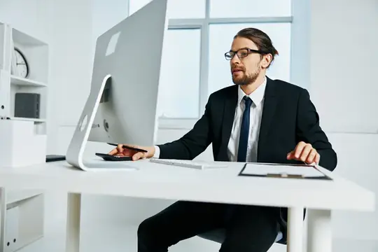 Мужчина в очках и деловом костюме работает за компьютером в светлом офисе