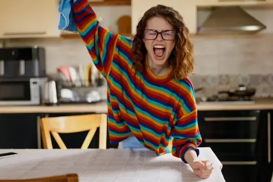 Кудрявая девушка в очках и радужном свитере весело танцует на кухне