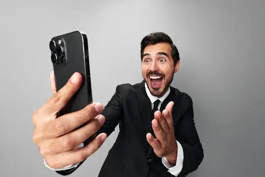 Мужчина-бизнесмен держит в руке телефон, удивлённо смотрит на экран во время видеозвонка, делает селфи и улыбается, обнажая зубы