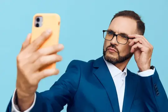 Мужчина в очках и синем пиджаке делает селфи на жёлтый смартфон