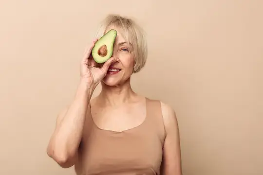 Улыбающаяся пожилая женщина с короткими волосами позирует с половинкой авокадо возле лица на нейтральном фоне