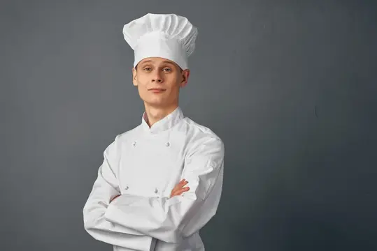 Молодой повар в белом колпаке и кителе со скрещёнными руками