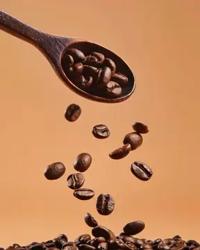 Кофейные зёрна падают из деревянной ложки на бежевом фоне