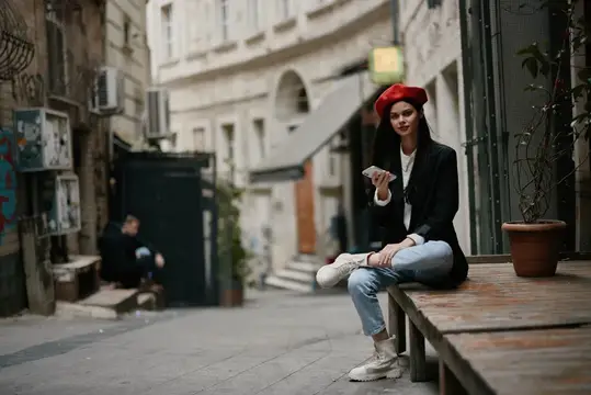 Стильная женщина-туристка в стильной одежде, в куртке и красном берете, сидит на улице с телефоном в руке, мобильный телефон во время путешествия, французский стиль, кинематографичный цвет