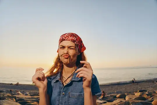 Игривая женщина, строящая забавную гримасу в красной бандане и джинсовой рубашке, стоит на каменистом пляже на закате, со спокойным морем и теплым золотистым светом на заднем плане, вызывая чувство…