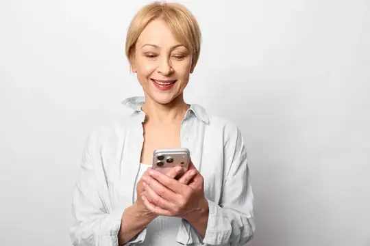 Улыбающаяся блондинка в белой рубашке смотрит в смартфон на светлом фоне