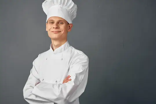 Молодой повар в белом колпаке и кителе со скрещёнными руками