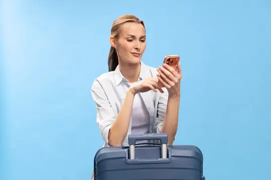 Светловолосая женщина с чемоданом смотрит в смартфон на голубом фоне