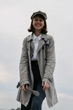 Улыбающаяся девушка в кепке и плаще на фоне пасмурного неба
