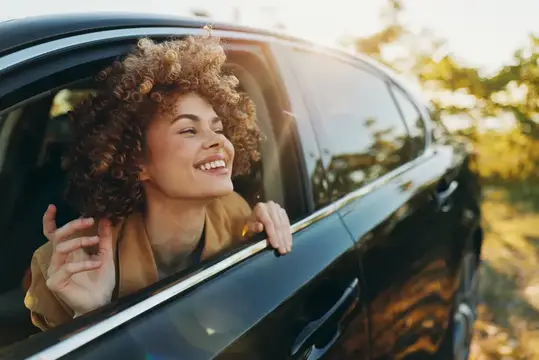Кудрявая девушка улыбается и машет рукой из окна автомобиля