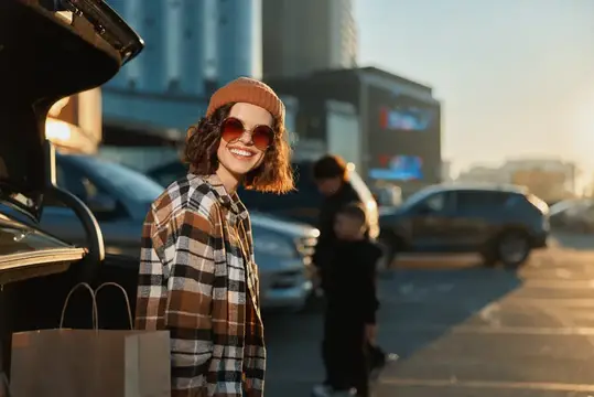 Улыбчивая девушка в клетчатой рубашке и очках у машины на закате