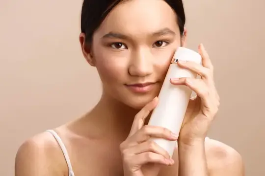 Азиатская девушка с белым флаконом у лица на бежевом фоне