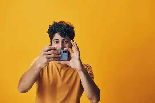 Кудрявый парень в жёлтой футболке фотографирует на смартфон на жёлтом фоне