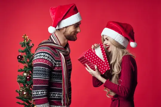 Пара в новогодних колпаках с подарком у ёлки на красном фоне