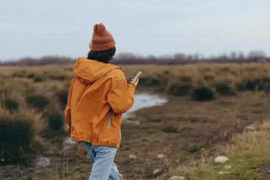 Человек в оранжевой куртке и шапке идёт с телефоном по осеннему лугу