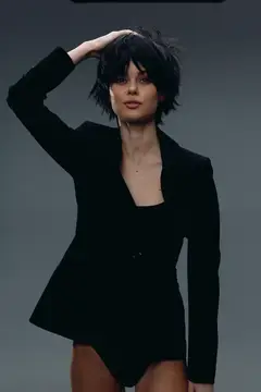 Стильная молодая женщина с короткими черными волосами уверенно позирует в черном пиджаке и боди на нейтральном сером фоне