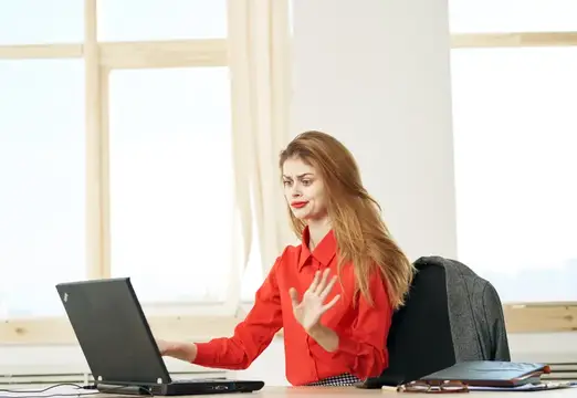 Рыжеволосая женщина в красной рубашке работает за ноутбуком в светлом офисе