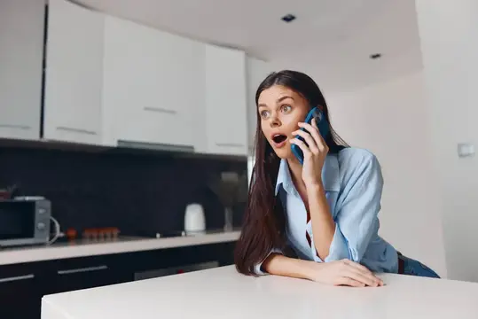 Удивлённая девушка с телефоном у стола на светлой кухне