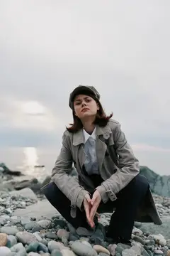Молодая женщина в кепке и плаще сидит на камнях у моря