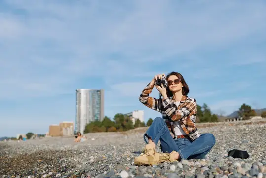 Девушка в клетчатой рубашке фотографирует на галечном пляже