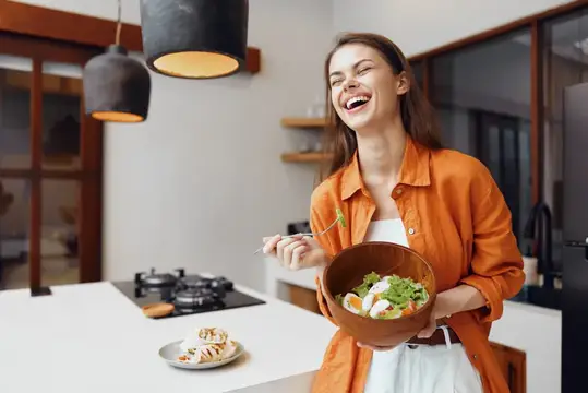 Смеющаяся девушка в оранжевой рубашке с салатом на кухне