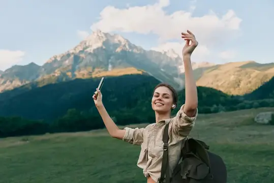 Улыбающаяся молодая женщина в повседневной рубашке и с рюкзаком стоит на открытом лугу на фоне гор, передавая ощущение приключения и свободы в ясный день