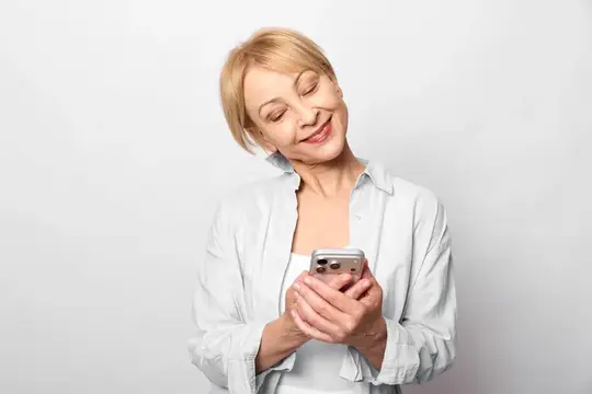 Пожилая женщина в белой рубашке держит смартфон, тихо смеется, в то время как технологии поддерживают обмен сообщениями для общения в социальных сетях