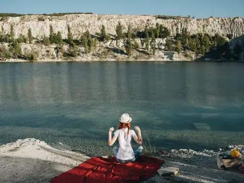 Женщина в белой футболке сидит на красной ткани возле моря на природе, на голове у неё шляпа