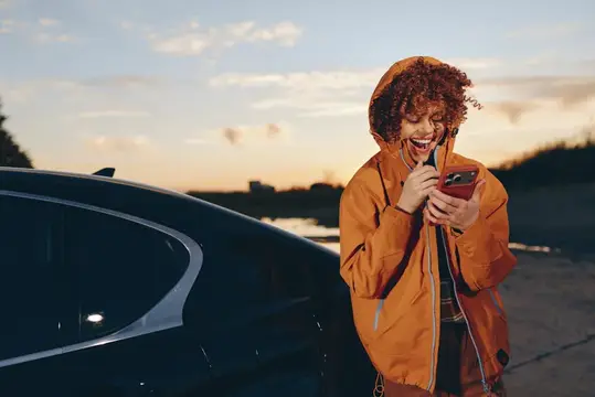 Рыжеволосая девушка в оранжевой куртке смеётся, глядя в телефон у машины на закате