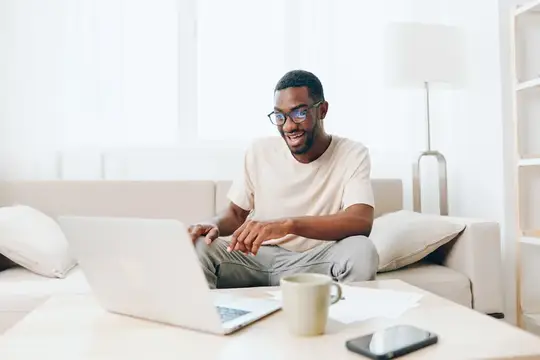 Улыбающийся темнокожий мужчина в очках работает за ноутбуком на диване