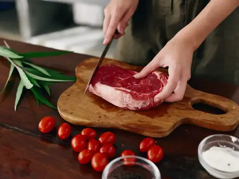 Женщина с ножом в руке нарезает свежее стейковое мясо для жарки на кухне; на столе соль, перец и другие специи, красные помидоры черри и зелень; она готовит ужин