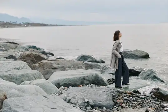 Молодая женщина в плаще стоит на камнях у моря в пасмурный день