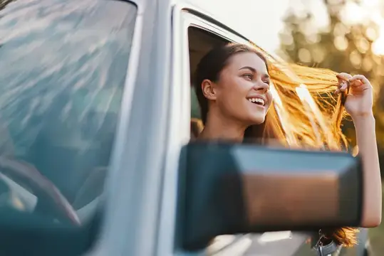 Улыбающаяся девушка высунулась из окна автомобиля и держит волосы на закате