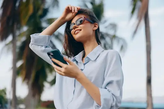 Улыбающаяся азиатская девушка в солнцезащитных очках со смартфоном на фоне пальм